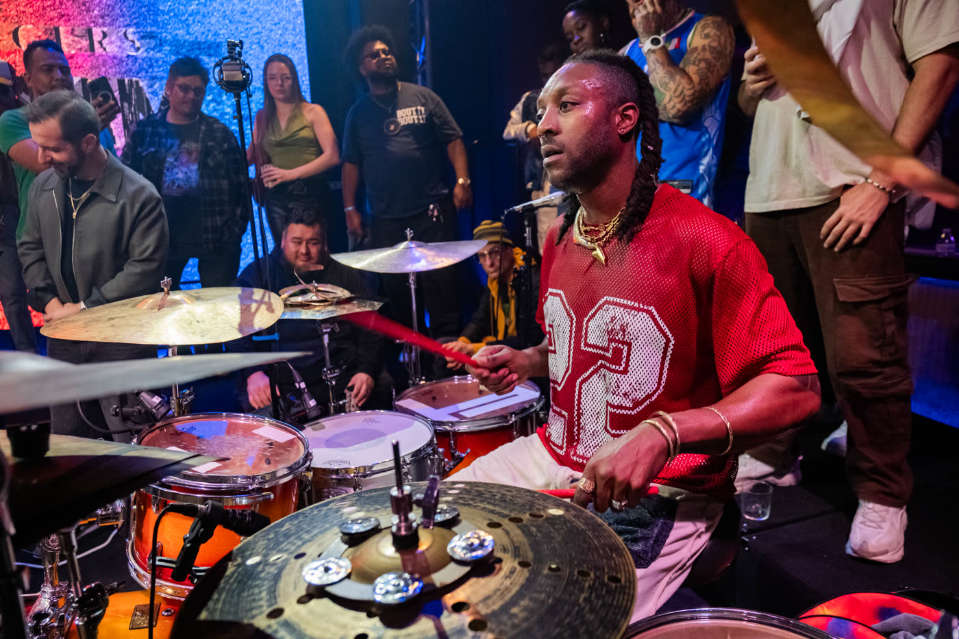 A drummer in a red jersey playing while the crowd stands close behind the kit.
